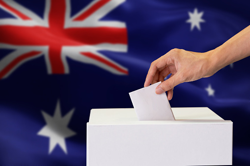 Close-up of human hand casting and inserting a vote and choosing and making a decision what he wants in polling box with Australia flag blended in background